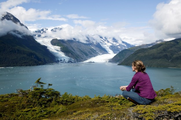 a woman crouches and looks out over the sea with a glacier in the background