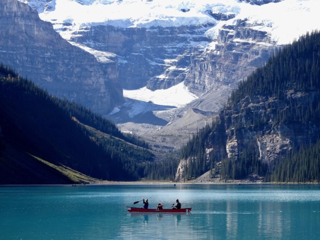 three people paddle a canoe on the turquoise water of Lake Louise with mountains in the background