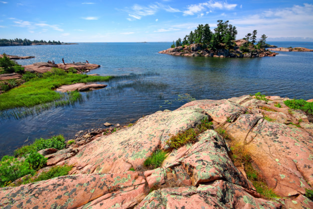 A scenic view of a rugged coastline in Ontario, Canada, with pink and green rocky terrain leading to a clear blue lake, small islands with trees in the distance, and a partly cloudy sky.