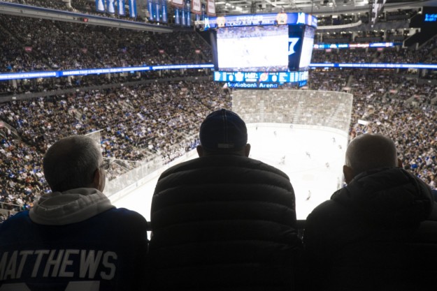 three men watch ice hockey in a crowded stadium