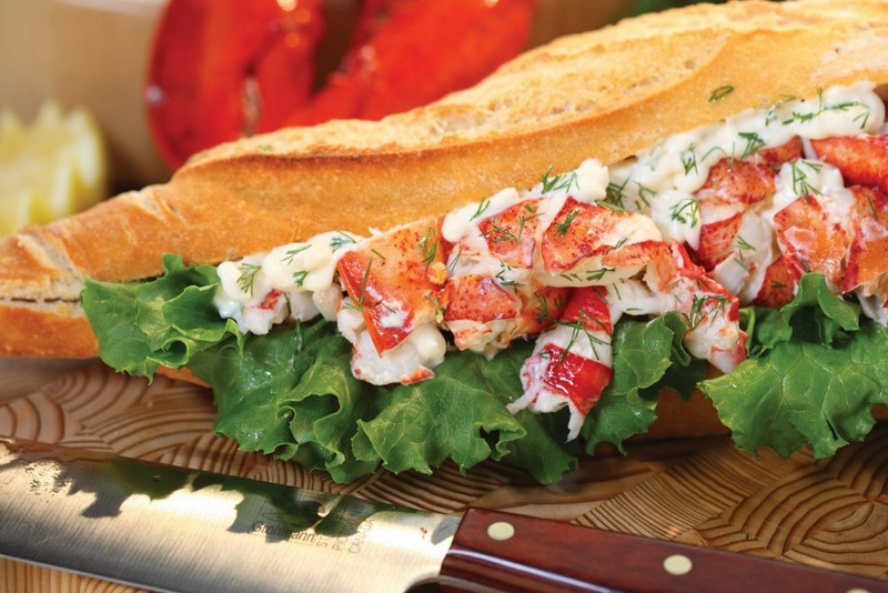 A lobster roll sandwich with chunks of lobster meat from the Atlantic Canada provinces, mayo, and dill in a French baguette, garnished with fresh lettuce, served on a wooden board.
