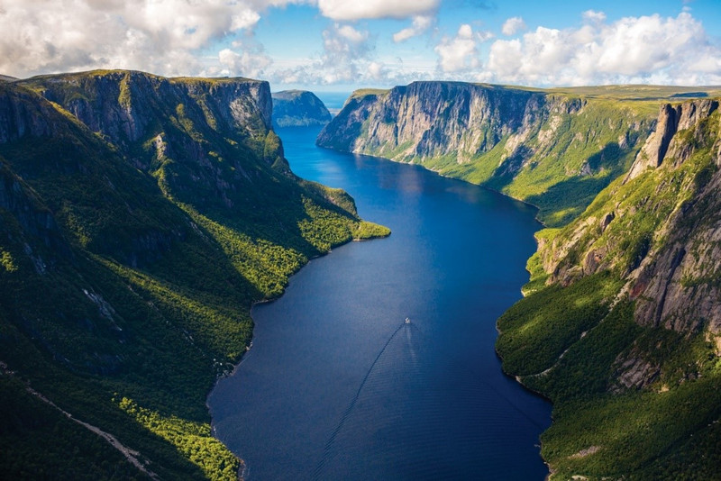 Aerial view of a scenic fjord in Nova Scotia, with steep, forest-covered cliffs on either side and a narrow river running through the center, under a bright blue sky with clouds.