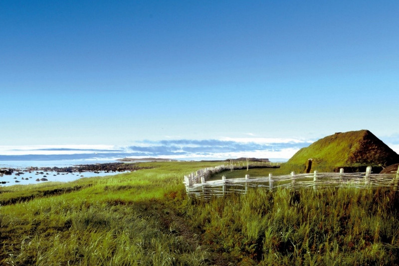 A panoramic view of a lush green coastal landscape in Nova Scotia with a traditional thatched-roof structure and a wooden fence extending towards the sea under a clear blue sky.
