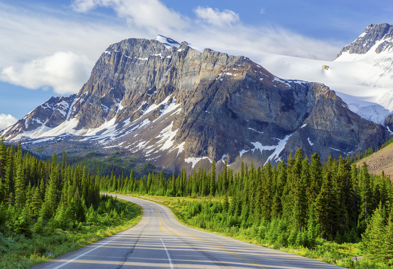 A scenic road, one of the premier routes in Canada, winds through a lush forest towards a towering snow-capped mountain under a bright blue sky.