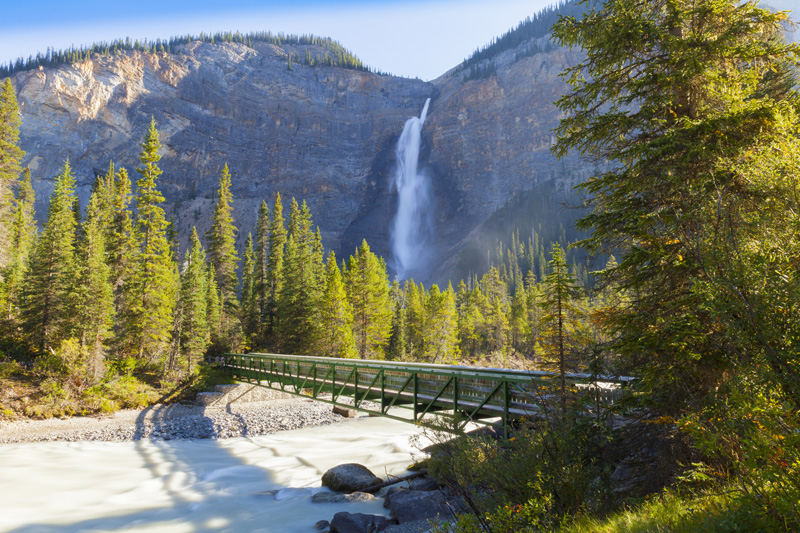 A scenic view featuring a tall, single-drop waterfall cascading into a river, surrounded by dense evergreen trees under a clear blue sky. A metal bridge crosses the river, enhancing the sights and attractions