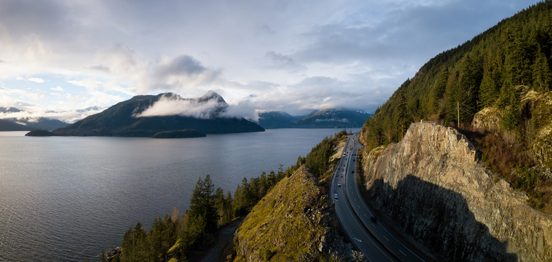 Panoramic view of a scenic coastal road curving along a rugged cliff with mountains shrouded in clouds across the water, captured during early evening light in Canada.