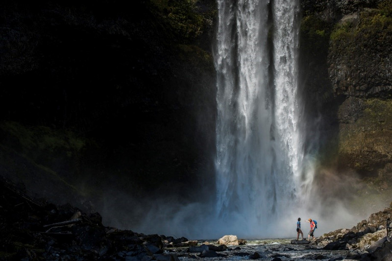 Two hikers stand near the base of a tall waterfall in Canada, with mist rising around them and rugged rocks scattered nearby, highlighting the scale and power of the natural scene.