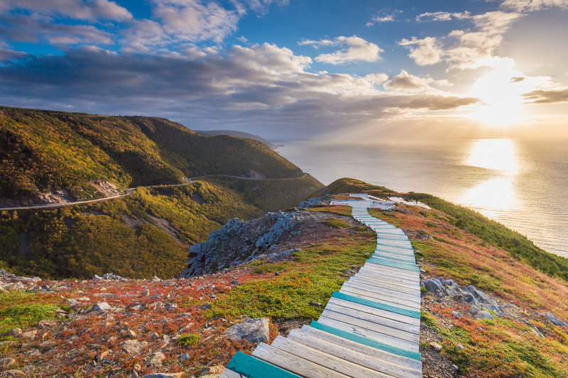 A scenic landscape featuring a wooden boardwalk meandering through colorful, rugged terrain under a vibrant sunset sky, with the ocean extending into the horizon, is one of Canada's standout sights and attractions.