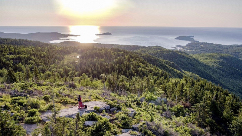 Aerial view of a person meditating on a rocky outcropping overlooking a lush forest, islands, and a sunset over the standout routes of Canada drives.