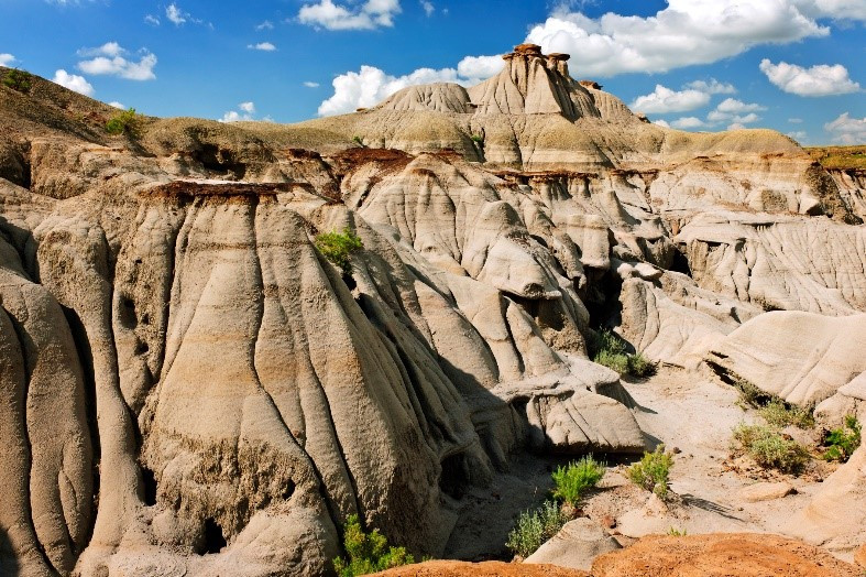 A landscape of rugged badlands with eroded rock formations, standout routes, and sparse green vegetation under a bright blue sky with puffy clouds.