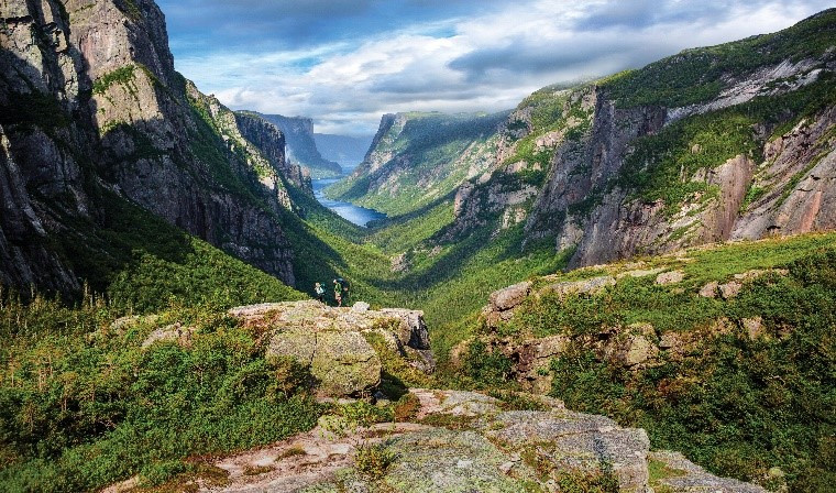 A scenic overlook of a deep valley with steep cliffs, featuring lush greenery and a distant lake, under a partly cloudy sky. Two people admire the view from a rocky outcrop along one of Canada