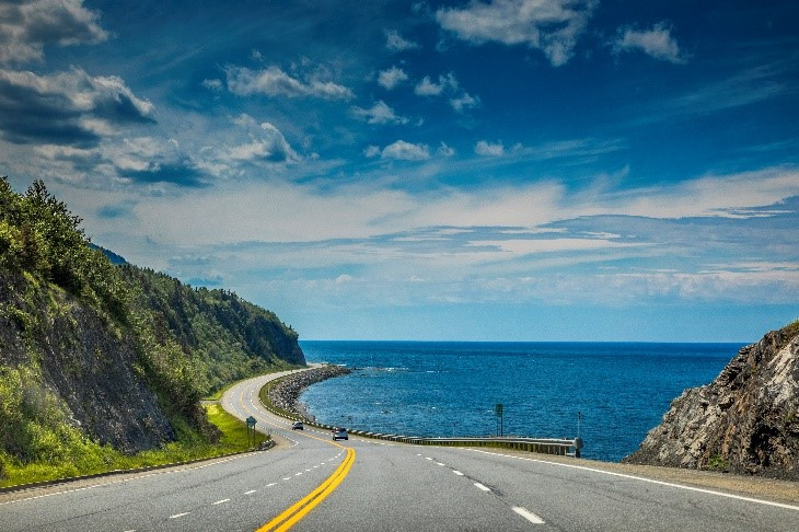 A picturesque coastal road, one of Canada's standout routes, curves beside a rocky cliff on one side and the bright blue sea on the other under a partly cloudy sky.