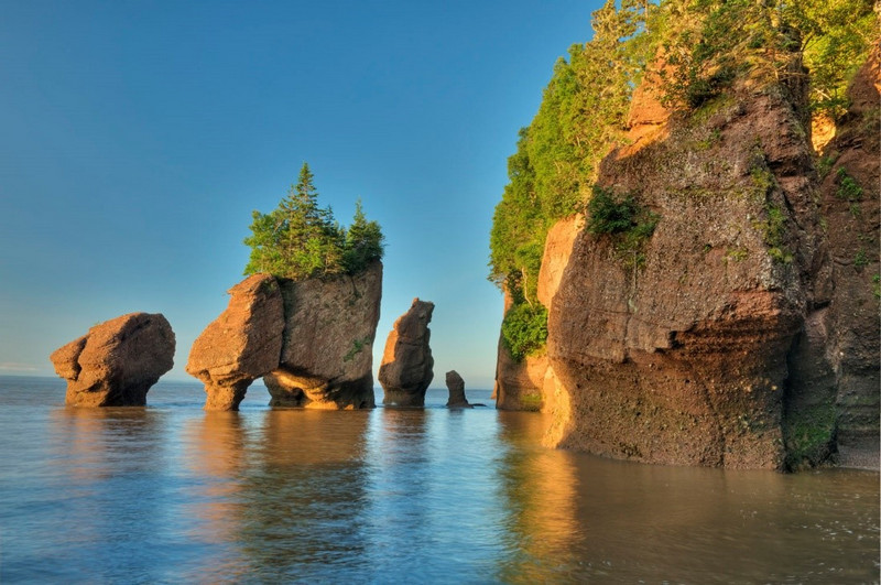 Rock formations resembling pillars stand in shallow waters near a forested cliff, illuminated by warm sunlight during a clear day, forming one of the standout sights and attractions in the area.