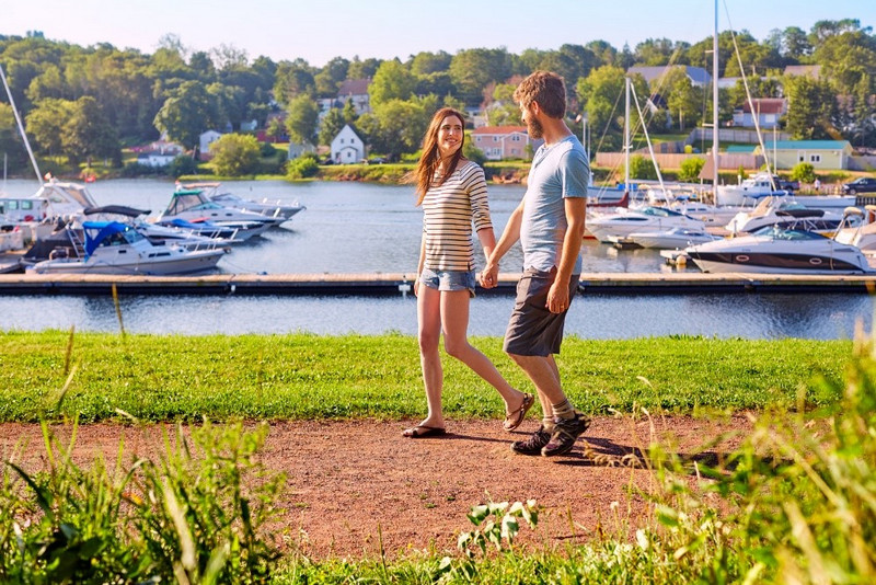 couple walks along a sandy trail with a boat marina behind them