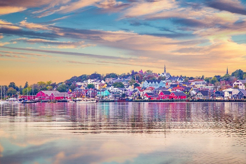 A picturesque waterfront town in Newfoundland and Labrador at sunset, featuring vibrant, colorful buildings and a calm bay reflecting the sky and townscape.